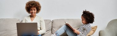 A joyful African American mother and daughter engage with a laptop while sitting on a cozy couch at home. clipart