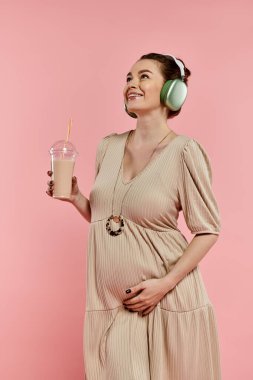 Kulaklık takan hamile bir kadın canlı pembe arka planda içkisini yudumluyor..