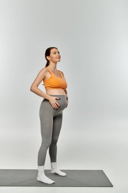 Aktif çalışan hamile bir kadın gri arka planda yoga minderinin üzerinde duruyor..