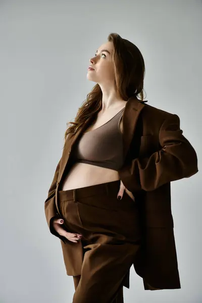 stock image A young pregnant woman elegantly poses in a stylish brown suit with a blazer against a grey background.