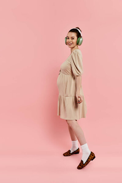 Беременная женщина в платье с радостью слушает музыку через наушники на розовом фоне.