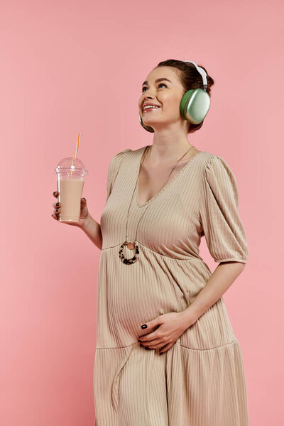 A young pregnant woman in a dress wearing headphones sips on a drink in a vibrant pink background.