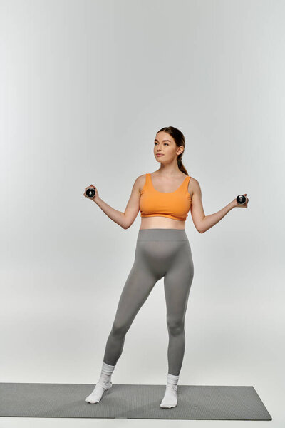 A pregnant woman in sportswear stands on a yoga mat, holding dumbbells in a balanced pose on a grey background.