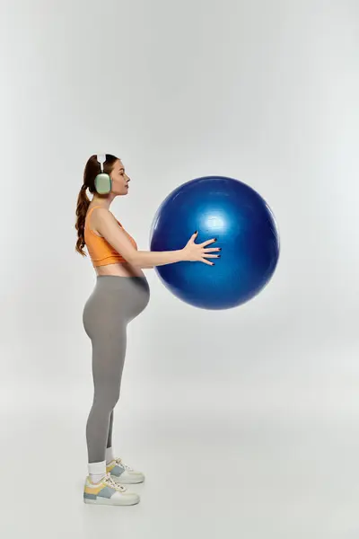 stock image A young, sporty pregnant woman in activewear holds a large blue ball on a grey background.
