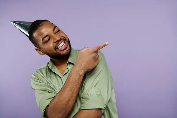 stock image A young African American man wearing braces pointing enthusiastically while donning a festive party hat against a purple backdrop.