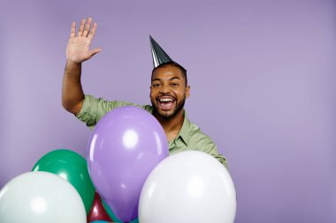 Young African American man in party hat smiling, holding colorful balloons against a purple background. clipart