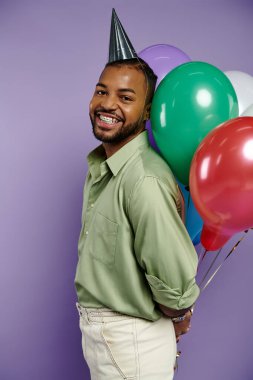 Young African American man in a party hat happily holds colorful balloons on a purple background. clipart