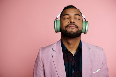 A young African American man listening to music through headphones on a pink background. clipart