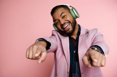Genç Afro-Amerikalı adam, parlak pembe arka planda kulaklık takarken kameraya işaret ediyor..