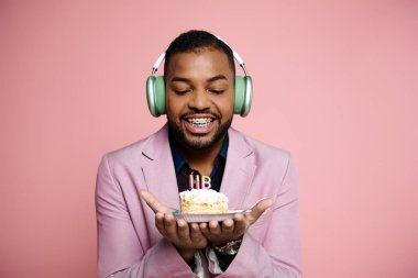 Telli neşeli genç Afrikalı Amerikalı adam pembe arka planda pasta tutarken kulaklıkla müzik dinliyor..