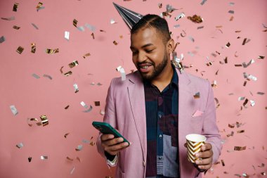 Young African American man in braces, happy, holding cup and cell phone, standing in party hat on a pink background. clipart