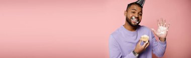 Mor gömlekli, diş teli takmış, pembe arka planda pastanın tadını çıkaran mutlu, Afro-Amerikalı bir adam..