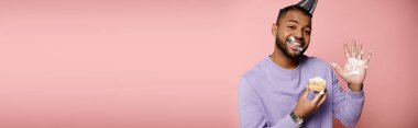 A happy, young African American man with braces, in a purple shirt, savoring a pastry on a pink background. clipart