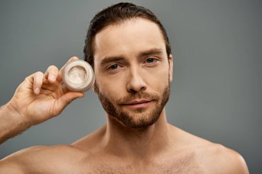 A shirtless, bearded man holding a jar of cream in his hand against a grey studio backdrop. clipart