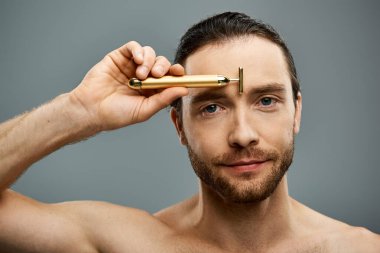 Shirtless man with beard touching golden razor to his forehead on a grey studio background. clipart