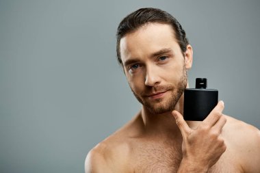 Gri arka planda siyah parfüm şişesi tutan üstsüz, sakallı bir adam..