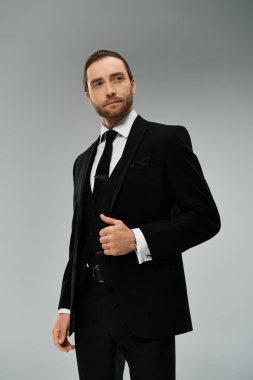 Takım elbiseli ve kravatlı yakışıklı, sakallı bir iş adamı gri bir arka plana karşı stüdyoda kendine güvenen bir poz veriyor..