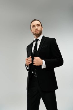 Takım elbiseli ve kravatlı şık, sakallı bir iş adamı gri stüdyo arkaplanına karşı kendine güvenen bir poz veriyor..