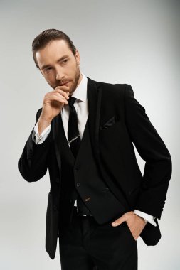 Yakışıklı ve sakallı bir iş adamı, şık bir smokinin içinde, gri bir arkaplana karşı kendine güvenen bir şekilde poz verir..