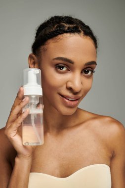 Askılı bluzlu genç Afro-Amerikalı kadın, gri arka planda bir şişe sıvı tutuyor..