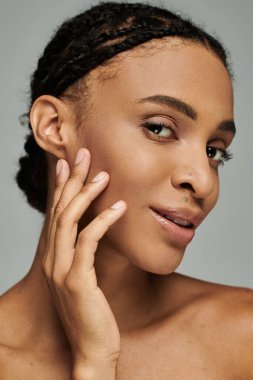 Saçları örgülü genç bir Afro-Amerikalı kadın. Gri bir arka planda cildiyle ilgileniyor..