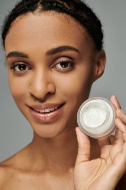 Genç Afro-Amerikalı kadın, askısız üst tutucu krem kavanozunda, cilt rutinine odaklanmış, gri arka planda....