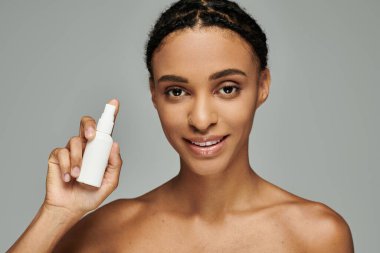 Young African American woman in strapless top holds lotion bottle, prioritizing skin care routine on grey background. clipart