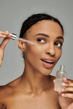 Üstü çıplak genç Afrikalı Amerikalı kadın gri arka planda serum şişesi tutuyor..