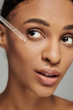 Askısız bluzlu genç bir Afrikalı Amerikalı kadın gri arka planda serum uyguluyor..
