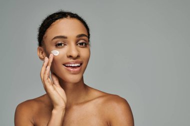 Askısız üst kısmı olan genç Afrikalı Amerikalı kadın beyaz bir arkaplan ile beyaz yanağına nazikçe cilt kremi sürüyor..