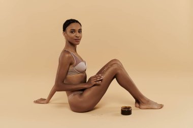 Güzel, zayıf, bikinili Afro-Amerikan bir kadın yerde oturuyor, sakin bir ortamda vücuduna bakıyor..