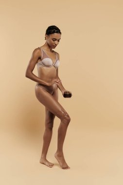 İnce, güzel bir Afro-Amerikan kadın beyaz sutyen ve bronz külot giyiyor, bej bir arka planda vücuduna bakıyor..