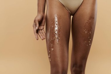 African American woman showcasing her beauty in a bikini coated with sugar scrub on legs on a beige backdrop. clipart