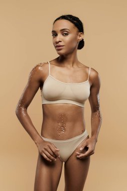 A pretty, slim African American woman is seen taking care of her body in tan lingerie on a beige background. clipart