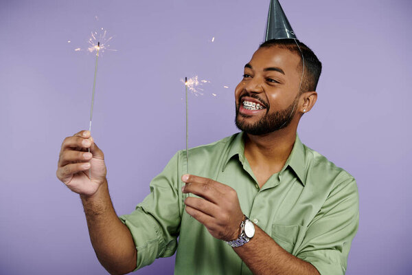 A young African American man in braces joyfully holding sparklers while wearing a party hat on a purple background.