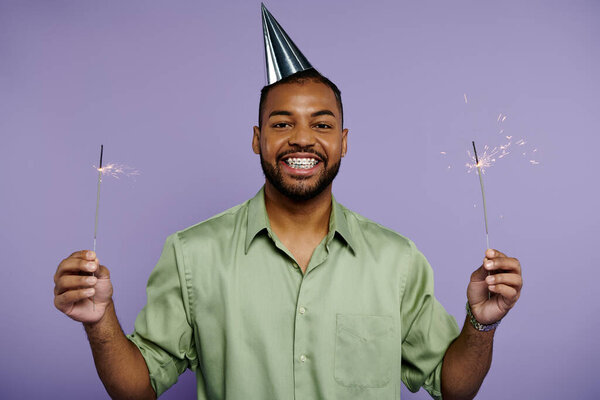 Young black man in braces smiling, holding two sparklers with a party hat on a purple background.