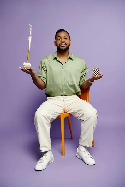 stock image A young African American man with braces smiles happily while sitting on a chair and holding a birthday cake on a purple background.