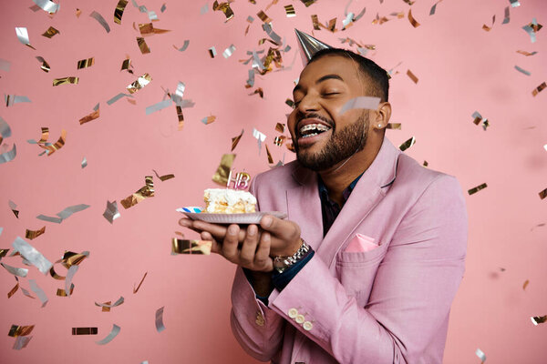 Young African American man in a pink suit happily holds a cake and confetti on a vibrant pink background.