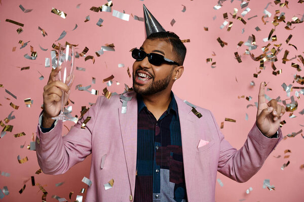 A young African American man in braces, looking happy, wears a party hat and sunglasses on a pink background.