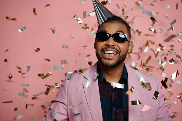 Young African American man joyfully wearing a party hat and sunglasses, exuding happiness on a pink background.