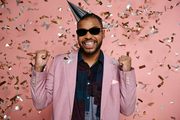 Young African American man beams in a party hat and sunglasses on a bright pink backdrop.
