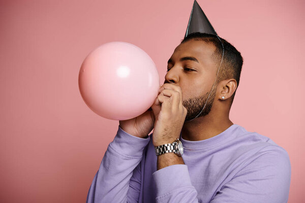 Young African American man wearing braces is happily blowing up a balloon while wearing a colorful party hat.