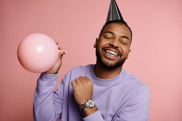 Young African American man with braces smiling and holding a balloon, wearing a colorful party hat on a pink background.