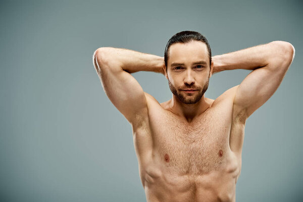 A shirtless man with his hands on his head, showcasing strength and vulnerability in a grey studio setting.