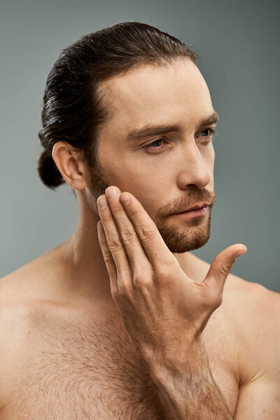 Handsome shirtless man with a beard, deep in thought, resting his hand on his face against a grey studio backdrop.