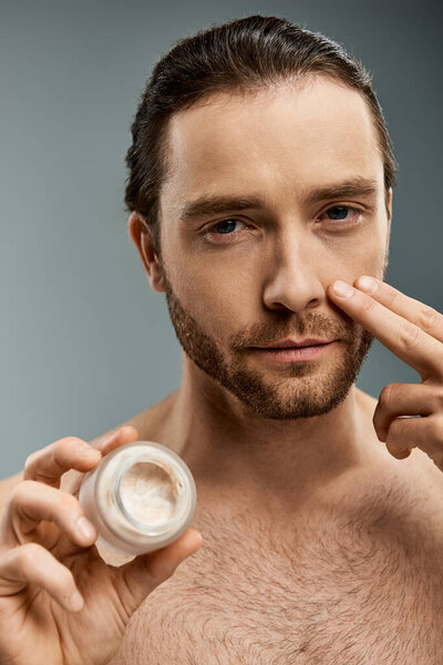 Handsome shirtless man with a beard holding a jar of cream against a grey backdrop.