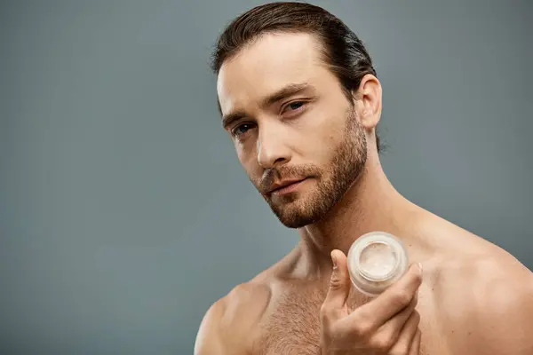 stock image A handsome, shirtless man with a beard holding cream in his hand against a grey studio backdrop.