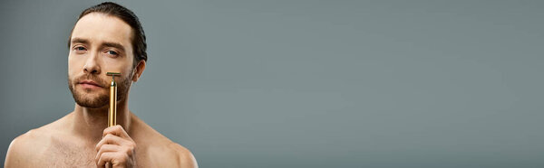 Shirtless man with beard holding golden razor against grey background.