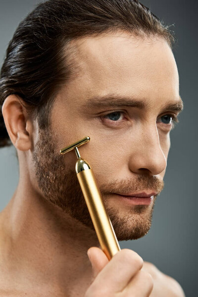Shirtless bearded man carefully holds a golden razor in his hand against a grey studio backdrop.