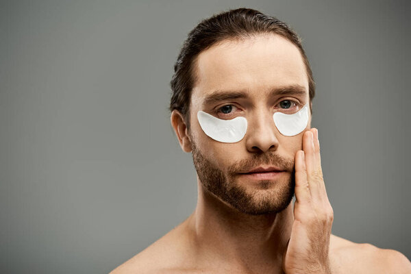 A handsome and shirtless man with a beard wearing eye patches on his face in a studio against a grey background.